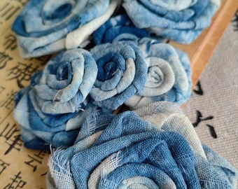 Pode incluir: Close-up de várias rosas de tecido feitas à mão em tons de azul e branco. As rosas são feitas de um tecido texturizado, com cada pétala cuidadosamente enrolada para criar uma forma de flor realista. A disposição está em um fundo neutro.