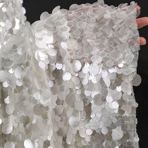 May include: A white fabric featuring a dense layer of overlapping circular sequins attached to a fine mesh base. The sequins are semi-translucent and reflective, creating a textured surface that covers the underlying white netting material.