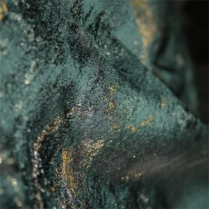 May include: Close-up of a textured fabric in shades of teal and dark green, with gold metallic accents. The fabric has a subtle sheen and a slightly crumpled appearance, suggesting a luxurious textile.