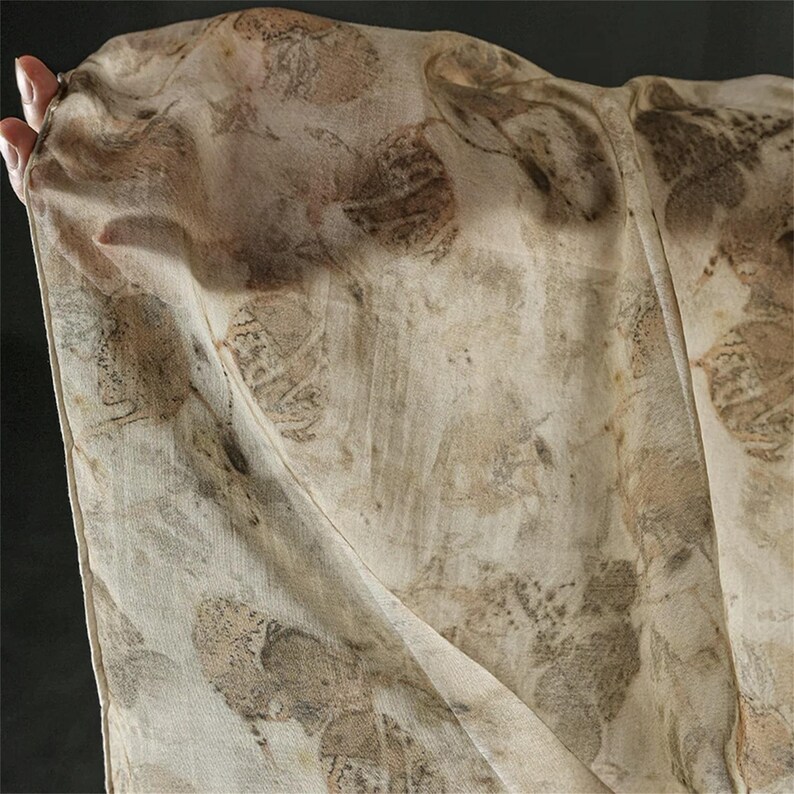 May include: A lightweight, semi-sheer scarf in a light beige color featuring a repeating botanical print of dark brown and tan leaves. The fabric has a soft, textured appearance with visible leaf vein details scattered across the entire surface.