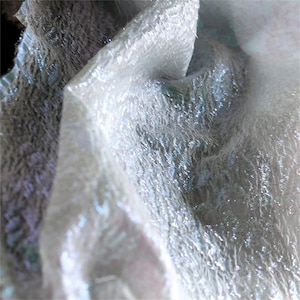 Può includere: Primo piano di un tessuto strutturato e iridescente. Il materiale sembra essere un bianco traslucido con sottili sfumature di blu e viola. Il tessuto è piegato, creando ombre e mettendo in risalto la trama.
