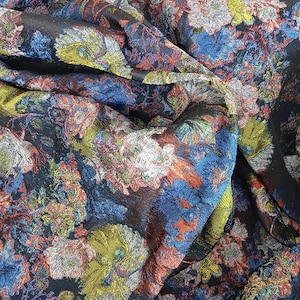 Op de afbeelding: Een stof met een dicht bloemenpatroon op een zwarte ondergrond. Het ontwerp bevat geweven draden in levendige tinten blauw, metallic zilver, helder limoengroen en oranje, wat zorgt voor een getextureerd en veelkleurig uiterlijk.