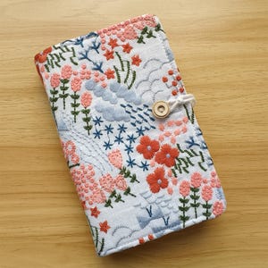 May include: Embroidered journal cover with a floral design in shades of coral, blue, and green on a white background. The cover features a button closure and is made of fabric. The journal cover is placed on a wooden surface.