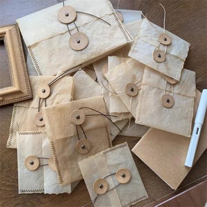 Puede incluir: Una colección de sobres de papel hechos a mano, cada uno sellado con botones de madera y cuerda. Los sobres son de color marrón claro, con un aspecto texturizado y envejecido. Los bordes están cosidos con hilo oscuro. También se ve un bolígrafo blanco y un pequeño marco de fotos.