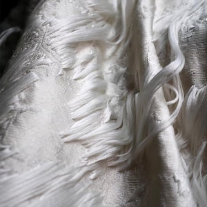 May include: Close-up of a white textile with a textured surface and long, flowing strands. The fabric appears soft and delicate, with a subtle sheen. The design features a combination of smooth and fringed elements.