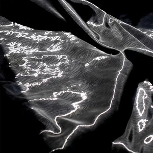 Può includere: Primo piano di un tessuto nero con un motivo strutturato e linee bianche irregolari. Il tessuto è drappeggiato, creando ombre e luci che enfatizzano la trama e il design. L'effetto complessivo è un mix di eleganza e arte astratta.