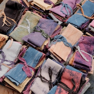 May include: A selection of small fabric bundles in various colours, secured with ribbons. The bundles showcase a range of shades, including blues, purples, pinks, and greens. The fabric edges are raw, giving a rustic appearance to the presentation.