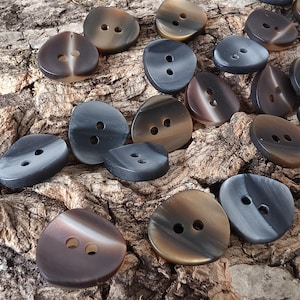 Puede incluir: Una colección de botones redondeados e irregulares con un acabado liso y pulido, presentando patrones de remolinos en tonos marrón oscuro, gris carbón y negro. Cada botón cuenta con dos orificios centrados para su fijación en prendas.