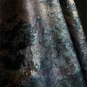 May include: Close-up of a textured fabric with a dark, abstract design. The fabric features a blend of deep blues, greens, and purples, accented with shimmering gold flecks. The overall effect is a rich, artistic textile.