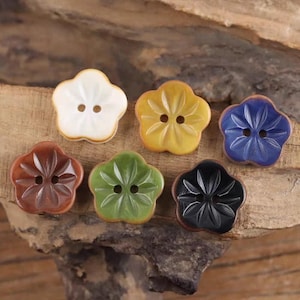 May include: A collection of seven flower-shaped buttons in various colors, including white, yellow, blue, brown, green, and black. Each button has two holes for sewing and a carved floral design. The buttons are displayed on a wooden surface.