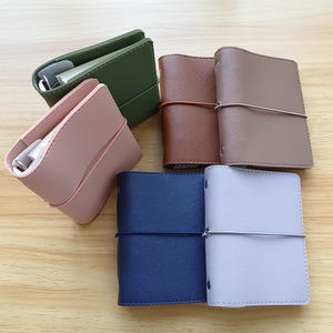 May include: A collection of leather journals and cases in various colors, including olive green, dusty rose, brown, taupe, navy blue, and lavender. The journals are bound with elastic closures, and the cases have a simple, minimalist design.