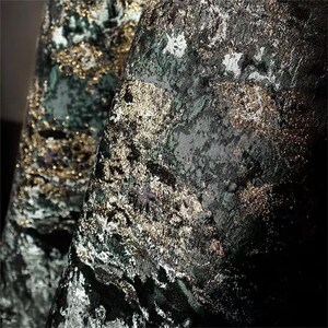May include: Close-up of a luxurious fabric with a textured, velvet-like appearance. The material features a dark green base with shimmering gold and silver accents, creating a rich, layered effect. The folds of the fabric add depth and dimension.