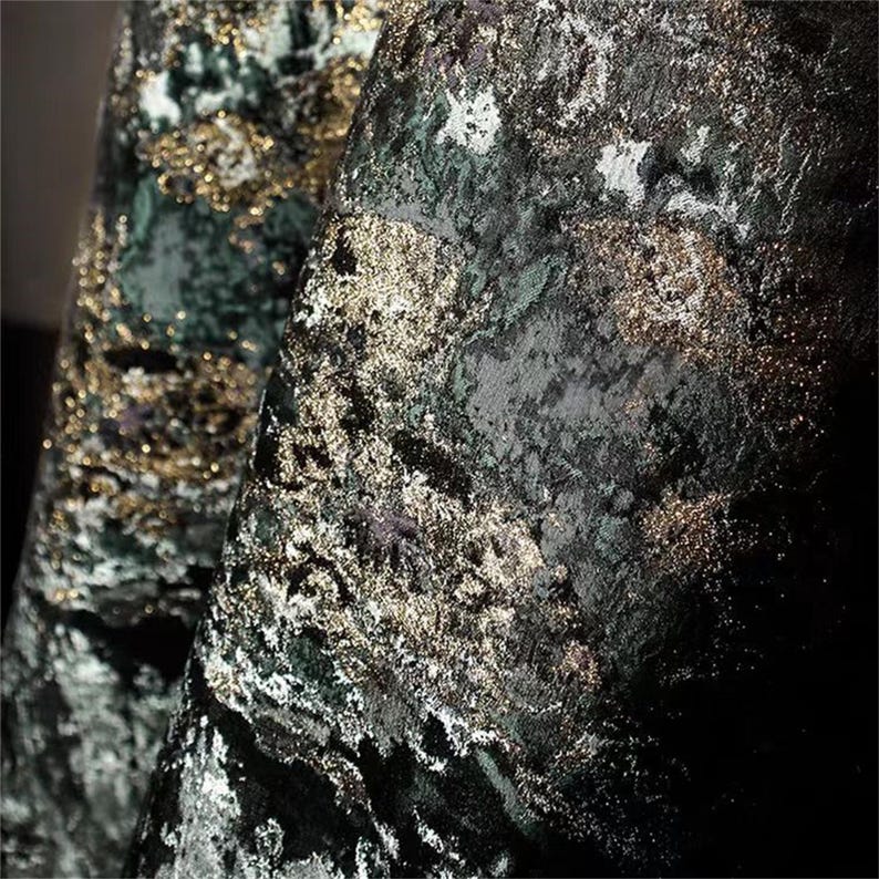 May include: Close-up of a luxurious fabric with a textured, velvet-like appearance. The material features a dark green base with shimmering gold and silver accents, creating a rich, layered effect. The folds of the fabric add depth and dimension.