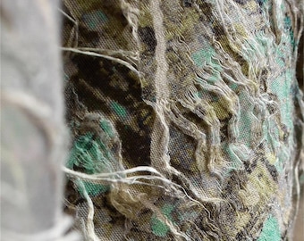 May include: Close-up of a textured fabric with a pattern of green, brown, and yellow. The fabric has a distressed appearance with frayed edges and white threads. The overall effect is a blend of colors and textures.