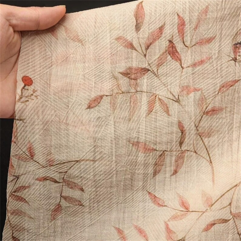 Op de afbeelding: Close-up van een beige stof met een bloemenpatroon. Het ontwerp toont roodbruine bladeren en stelen, tegen een subtiele rasterachtige achtergrond. De stof wordt door een hand vastgehouden.