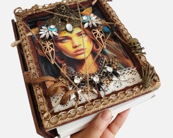 May include: A decorative journal featuring a portrait of a woman with a jeweled headdress. The journal is adorned with gold chains, a skull, and a hand-shaped embellishment. The cover is framed with a gold chain and a brown border.
