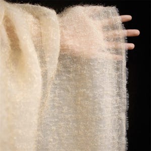 May include: A delicate, cream-coloured fabric with a textured appearance. The fabric is held up, showing its lightweight and airy quality. The edges have a slightly frayed texture, adding to its ethereal quality. The background is black.