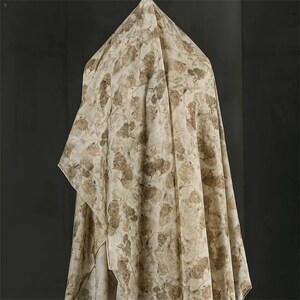 May include: A lightweight fabric drape featuring an all-over botanical print with scattered brown and tan leaf silhouettes on a cream-colored background. The material hangs in soft, vertical folds, displaying a repeating pattern of organic shapes.