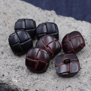 Puede incluir: Seis botones de cuero marrón oscuro con un diseño trenzado. Los botones cuadrados tienen bordes redondeados y un pequeño agujero para coser. Se muestran sobre una superficie gris rugosa. Cada botón mide aproximadamente 2 cm de diámetro.