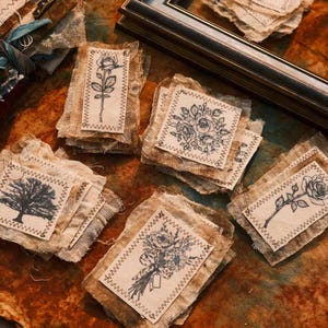 May include: A collection of rectangular fabric patches with frayed edges, each featuring a black ink illustration of botanical elements or a tree. The patches are stitched with a zigzag pattern onto a textured, light-coloured fabric base.