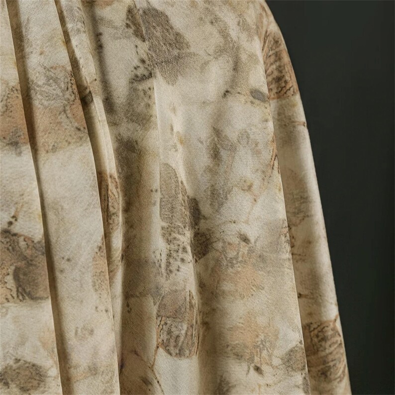 May include: A piece of fabric featuring a botanical print with various leaf shapes in shades of brown, tan, and cream. The material has a soft, draped texture with visible folds and pleats, showcasing an organic, earth-toned pattern throughout.