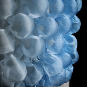 Può includere: Primo piano di un tessuto blu chiaro strutturato con un design a strati a forma di bolla. Il tessuto appare semitrasparente, con una sfumatura di colore dal blu chiaro al blu medio. Il design ricorda una forma floreale o organica.