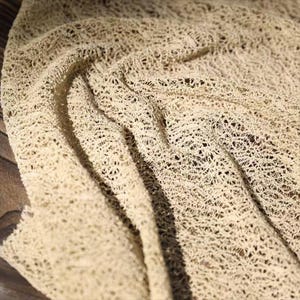May include: Close-up of a beige, open-weave fabric with a delicate, net-like texture. The fabric is draped, creating soft folds and shadows. The material appears to be lightweight and airy, suitable for various craft projects or apparel.