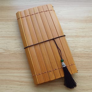 Pode incluir: Um caderno retangular de bambu com fecho de cordão castanho escuro e borla decorativa. O caderno é feito de finas ripas de bambu castanho claro presas com fio cor de cobre. A borla tem uma conta verde e uma tampa de metal prateado.