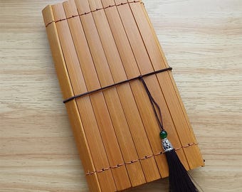 Puede incluir: Un cuaderno rectangular de bambú con cierre de cordón marrón oscuro y borla decorativa. El cuaderno está hecho de finas láminas de bambú marrón claro sujetas con hilo color cobre. La borla tiene una cuenta verde y una tapa de metal plateado.