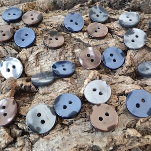 May include: A collection of round, two-hole buttons in various shades of blue, gray, and brown. Each button features a smooth, polished surface with subtle color variations, arranged in a scattered pattern across the frame.