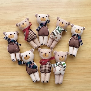 Könnte beinhalten: Eine Sammlung handgefertigter Teddybär-Ornamente. Jeder Bär ist beige mit einer braunen Nase und gestickten Details. Sie tragen gemusterte Schals und Outfits in verschiedenen Farben, darunter Braun, Rot und Grün. Die Bären sind auf einer hellen Holzoberfläche angeordnet.
