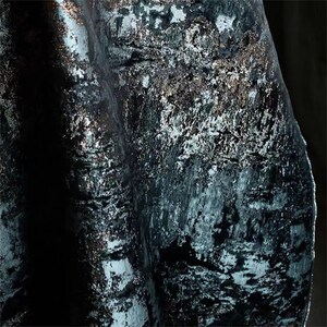 May include: Close-up of a dark teal fabric with a textured, metallic finish. The fabric has a velvet-like appearance with silver and bronze accents, creating a shimmering effect. The folds and shadows add depth to the material.