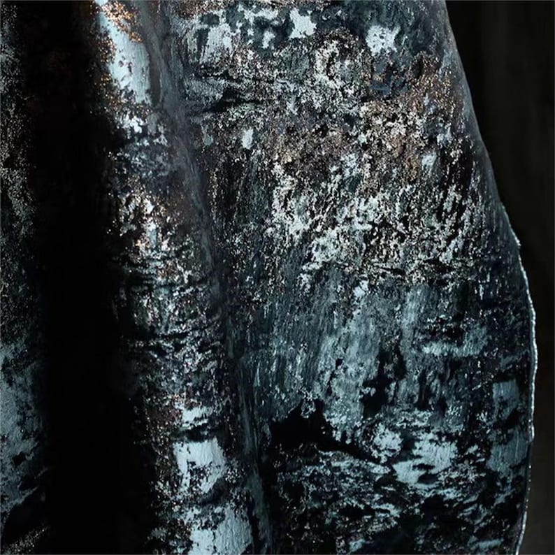 May include: Close-up of a dark teal fabric with a textured, metallic finish. The fabric has a velvet-like appearance with silver and bronze accents, creating a shimmering effect. The folds and shadows add depth to the material.