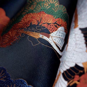 Puede incluir: Primer plano de una tela azul oscuro con bordados detallados. El diseño presenta una grulla, pinos y nubes en tonos rojos, verdes, blancos y dorados. La tela tiene una apariencia texturizada, lo que sugiere un textil de alta calidad.