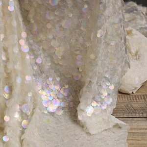 May include: A sheer white fabric featuring a dense overlay of circular, iridescent sequins. The sequins are semi-transparent with a pearlescent finish, reflecting light in various colors against the fine, lightweight mesh base material.