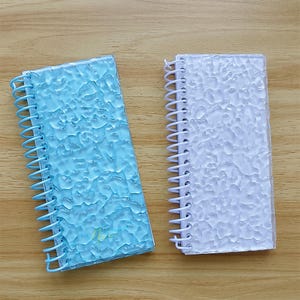 May include: Two spiral-bound notebooks with textured covers. One has a light blue cover and a matching blue spiral binding. The other has a white cover and a white spiral binding. Both notebooks are on a wooden surface.