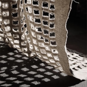 Peut inclure: Gros plan sur un tissu beige avec un motif quadrillé de découpes carrées. Le tissu est drapé, projetant une ombre avec un motif en losange sur une surface sombre. Les bords du tissu sont effilochés.