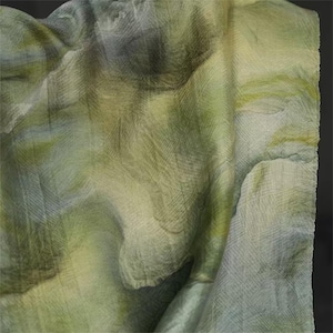 May include: A draped fabric with a watercolor-like effect. The textile features a blend of green, yellow, and gray hues, creating a soft, textured appearance. The folds and shadows add depth to the design, suggesting a lightweight material.