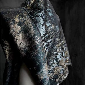 May include: Close-up of a dark fabric with a textured, abstract pattern. The fabric features a mix of black, teal, and gold tones, creating a shimmering effect. The material is draped, showcasing its flow and texture.