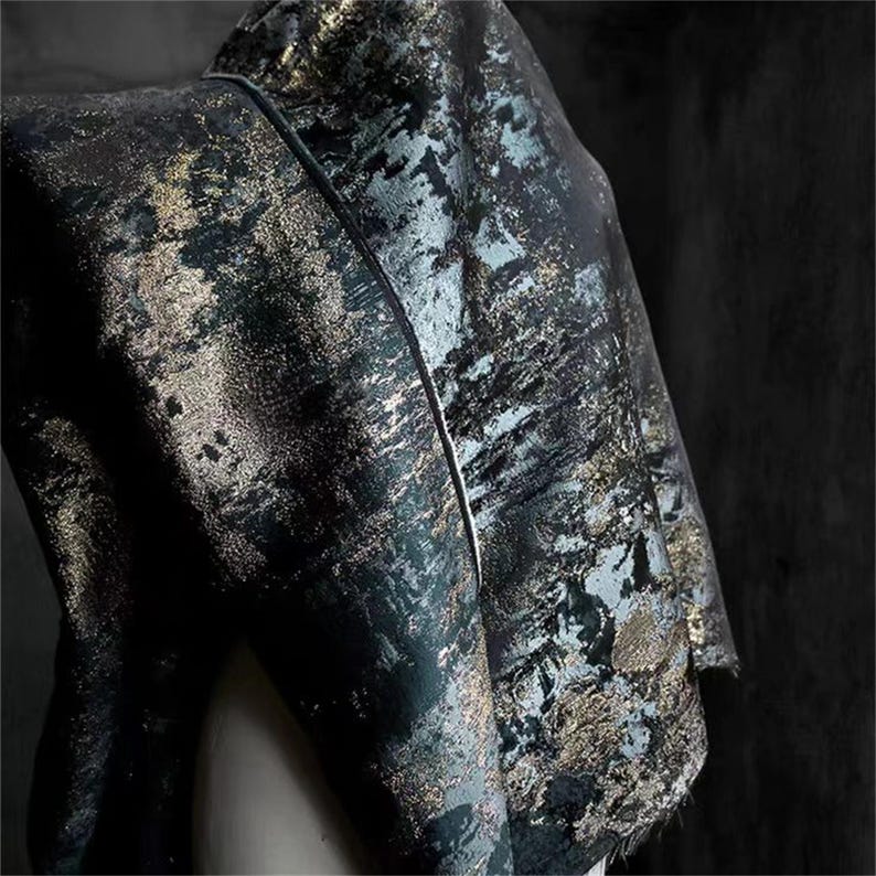 May include: Close-up of a dark fabric with a textured, abstract pattern. The fabric features a mix of black, teal, and gold tones, creating a shimmering effect. The material is draped, showcasing its flow and texture.