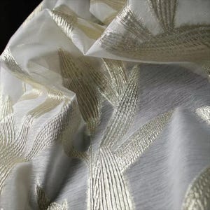 May include: A sheer white fabric featuring a repeating pattern of metallic gold embroidered leaf-like shapes. The material has a translucent quality with visible folds and gathers that create depth and texture across the surface of the textile.