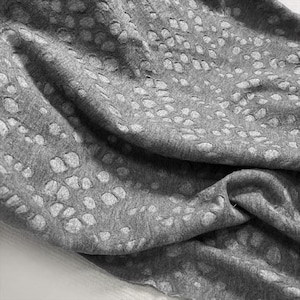 Puede incluir: Primer plano de una tela gris con un patrón de cortes circulares espaciados. La tela está drapeada, mostrando pliegues y sombras. El material parece suave y ligero, adecuado para ropa o decoración del hogar.