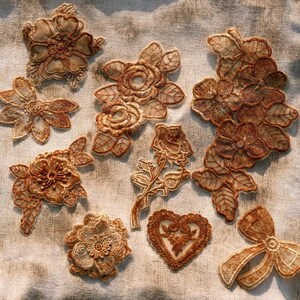 May include: A collection of intricately embroidered floral and bow appliques in shades of brown and tan. The appliques include various flower designs, a rose, and a heart-shaped piece, all with detailed stitching and delicate edges. These embellishments are ideal for crafting and textile projects.