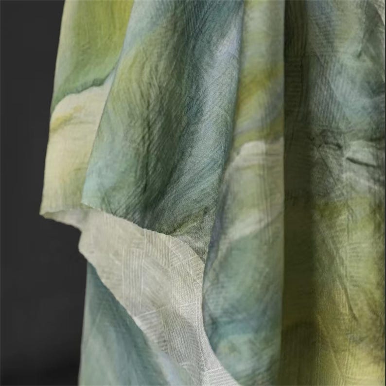 May include: Close-up of a flowing fabric with a watercolor effect. The textile features shades of green, blue, and yellow, creating a soft, blended appearance. The edge of the fabric has a white, textured border.