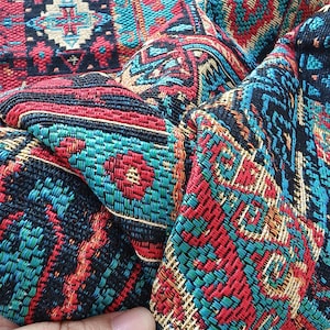 Pode incluir: Um tecido entrelaçado com um padrão geométrico denso e intrincado em tons vibrantes de vermelho, turquesa, preto e dourado. O material possui uma trama texturizada e resistente com motivos repetitivos que criam uma superfície complexa.