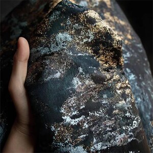 May include: Close-up of a dark fabric with a textured surface. The fabric has a black base with gold and silver metallic accents, creating a shimmering effect. The material is folded, revealing its layered texture and design.