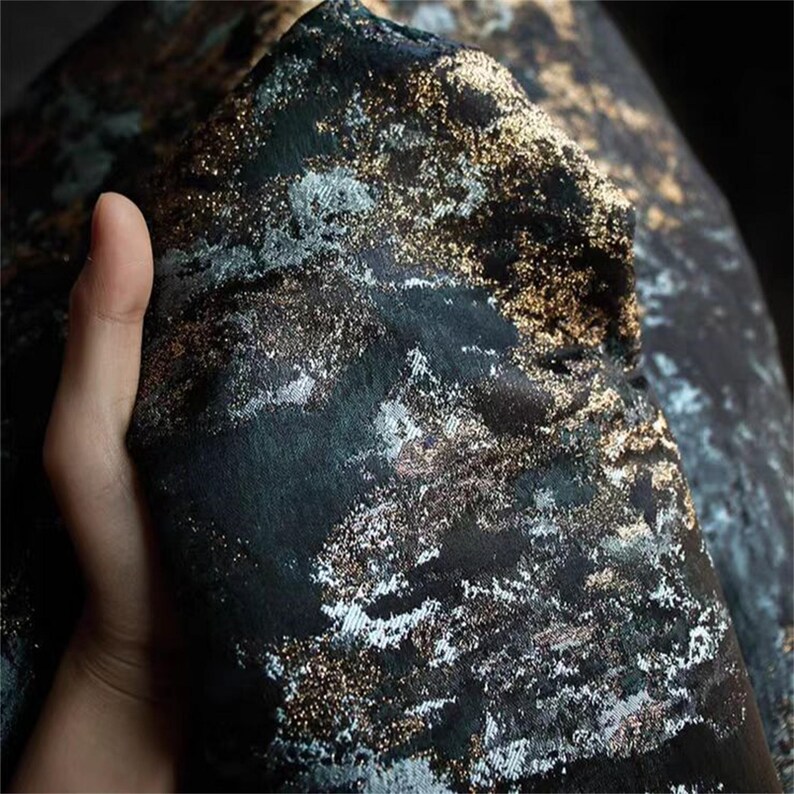 May include: Close-up of a dark fabric with a textured surface. The fabric has a black base with gold and silver metallic accents, creating a shimmering effect. The material is folded, revealing its layered texture and design.