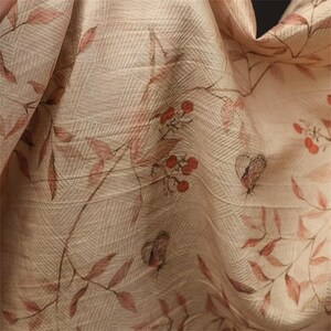 Op de afbeelding: Close-up van een beige stof met een botanische print. Het ontwerp toont rode bladeren, bessen en vlinders. De stof heeft een subtiel rasterpatroon, wat een textuur geeft. De algehele esthetiek is zacht en natuurlijk.