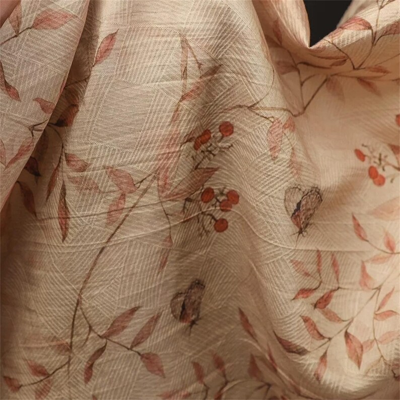 Op de afbeelding: Close-up van een beige stof met een botanische print. Het ontwerp toont rode bladeren, bessen en vlinders. De stof heeft een subtiel rasterpatroon, wat een textuur geeft. De algehele esthetiek is zacht en natuurlijk.
