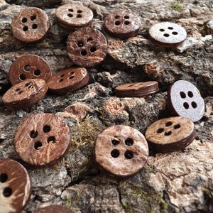May include: A collection of round, brown buttons with four holes each. The buttons feature a textured, natural surface with visible wood grain patterns and varying shades of dark and light brown, displaying a flat, circular shape with smooth edges.